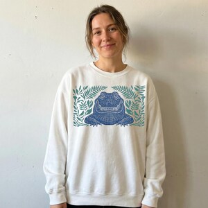 K&ouml;nnte beinhalten: Wei&szlig;es Sweatshirt mit einem blauen Alligatormotiv, das von t&uuml;rkisfarbenen Bl&auml;ttern umrahmt wird. Das Alligator hat detaillierte Schuppen und ist mittig auf der Vorderseite platziert. Der Text "Live Forever, Pal" ist auf dem Maul des Alligators aufgedruckt.