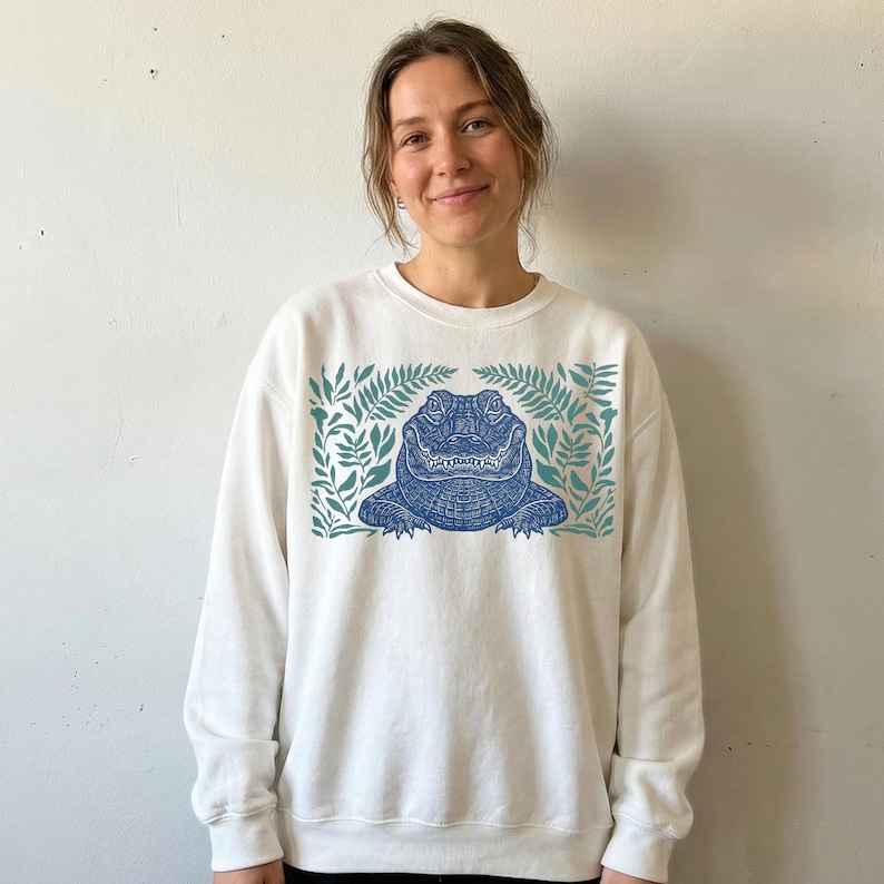K&ouml;nnte beinhalten: Wei&szlig;es Sweatshirt mit einem blauen Alligatormotiv, das von t&uuml;rkisfarbenen Bl&auml;ttern umrahmt wird. Das Alligator hat detaillierte Schuppen und ist mittig auf der Vorderseite platziert. Der Text "Live Forever, Pal" ist auf dem Maul des Alligators aufgedruckt.