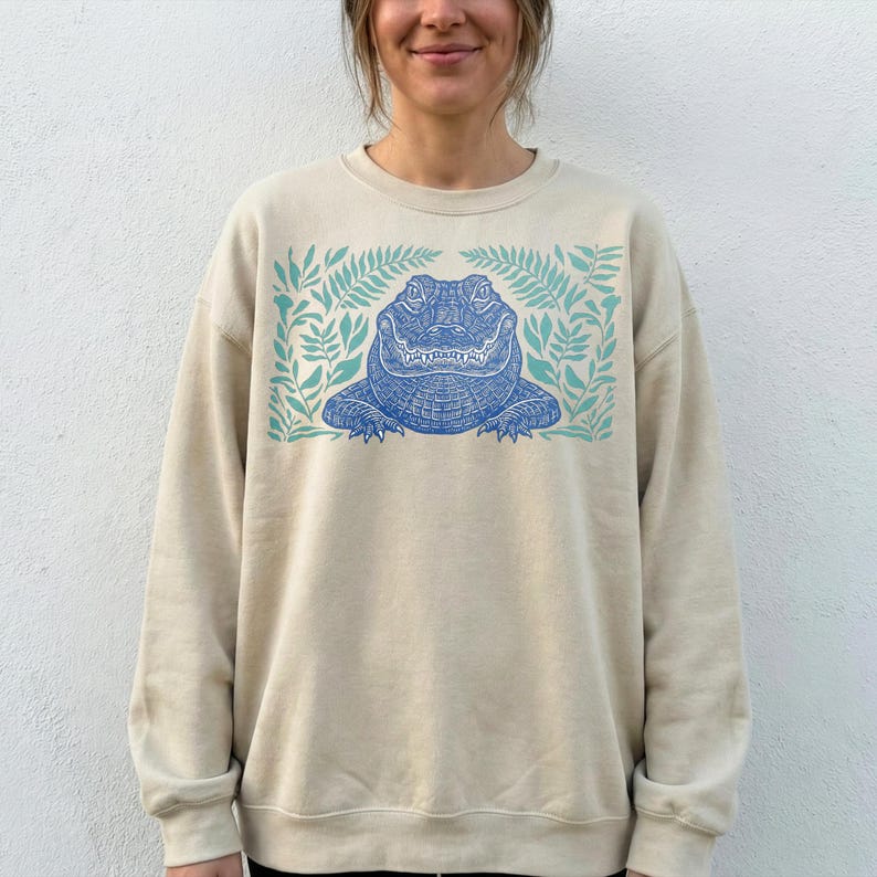 K&ouml;nnte beinhalten: Beiges Sweatshirt mit einem blauen Alligator-Design, umgeben von t&uuml;rkisfarbenen Farnbl&auml;ttern. Der Alligator hat detaillierte Schuppen und einen l&auml;chelnden Ausdruck. Das Sweatshirt hat einen Rundhalsausschnitt und lange &Auml;rmel.