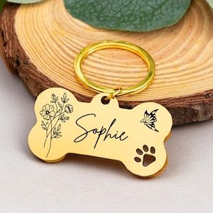 Könnte beinhalten: Ein goldfarbener, knochenförmiger Haustieranhänger mit dem Namen "Sophie" in Schreibschrift. Der Anhänger zeigt ein Blumendesign, einen Schmetterling und einen Pfotenabdruck. Er enthält einen goldfarbenen Schlüsselring.