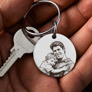 Peut inclure: Un porte-clés argenté avec un pendentif rond gravé d'une image détaillée d'une personne embrassant un enfant. Le porte-clés comprend une clé et un anneau métallique. L'image est tenue dans une main.