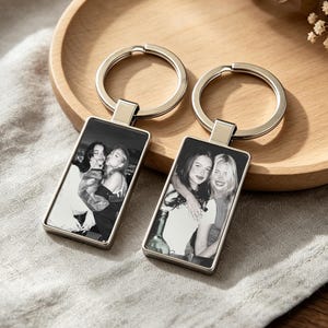 May include: Two rectangular metal keychains featuring black and white photographic prints of two people. Each keychain has a silver-toned frame and a circular split ring attachment, showcasing a clear, high-contrast image of two individuals.