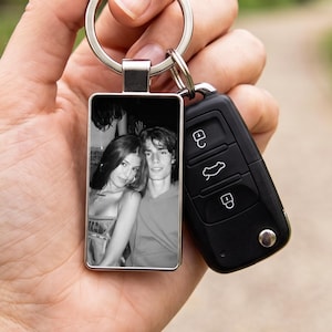 Puede incluir: Un llavero rectangular de metal con una fotografía en blanco y negro de dos personas. El llavero está unido a una anilla plateada, que también sostiene un mando de coche negro de plástico con tres botones y un botón metálico pequeño.