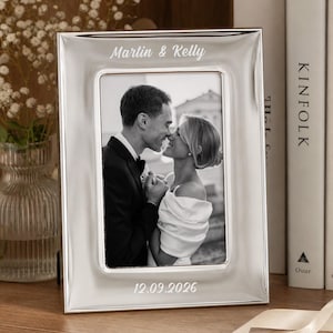 May include: A silver-toned rectangular photo frame featuring the engraved text Martin & Kelly at the top and 12.09.2026 at the bottom. The frame holds a black and white photograph of a couple embracing, with a smooth, polished metallic finish.