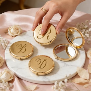 May include: Gold compact mirrors with engraved initials. One mirror is open, showing a reflective surface. The initials "R", "B", and "D" are displayed on the compacts. The mirrors are arranged on a white marble surface, surrounded by flowers.