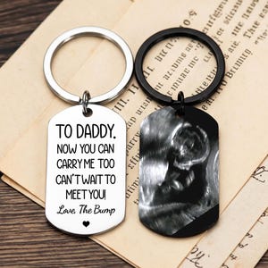 Op de afbeelding: Twee sleutelhangers in de vorm van een dogtag met ronde ringen. Eén zilverkleurige hanger bevat de zwarte tekst: TO DADDY. NOW YOU CAN CARRY ME TOO CAN'T WAIT TO MEET YOU! Love, The Bump. De andere zwarte hanger toont een grijstinten echofoto.
