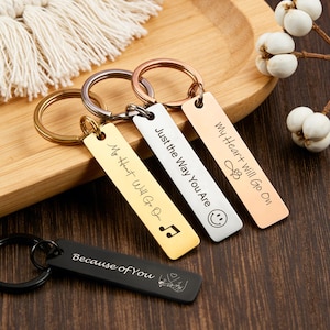 Op de afbeelding: Een verzameling sleutelhangers in goud, zilver en roségoud. Elke rechthoekige tag is gegraveerd met een andere boodschap, waaronder "My Heart Will Go On" en "Just the Way You Are". Eén zwarte sleutelhanger leest "Because of You".
