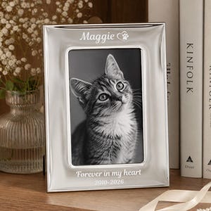 May include: A silver-toned rectangular metal picture frame featuring the engraved name Maggie with a paw print icon at the top and the text Forever in my heart 2010-2026 at the bottom, containing a black and white photograph of a tabby cat.