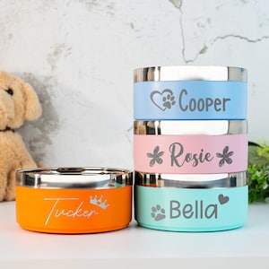 Puede incluir: Pila de cuatro cuencos coloridos para mascotas con bordes de acero inoxidable. Los cuencos son de color naranja, rosa, azul claro y turquesa. Cada cuenco tiene grabado el nombre de una mascota, junto con una huella de pata, un coraz&oacute;n o un dise&ntilde;o de corona. Un perro de peluche est&aacute; en el fondo.