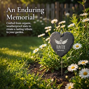 May include: A heart-shaped, dark gray slate memorial marker with the name "Hunter" and dates 2009-2026, featuring a paw print with wings design. The marker is in a garden with daisies and green grass.