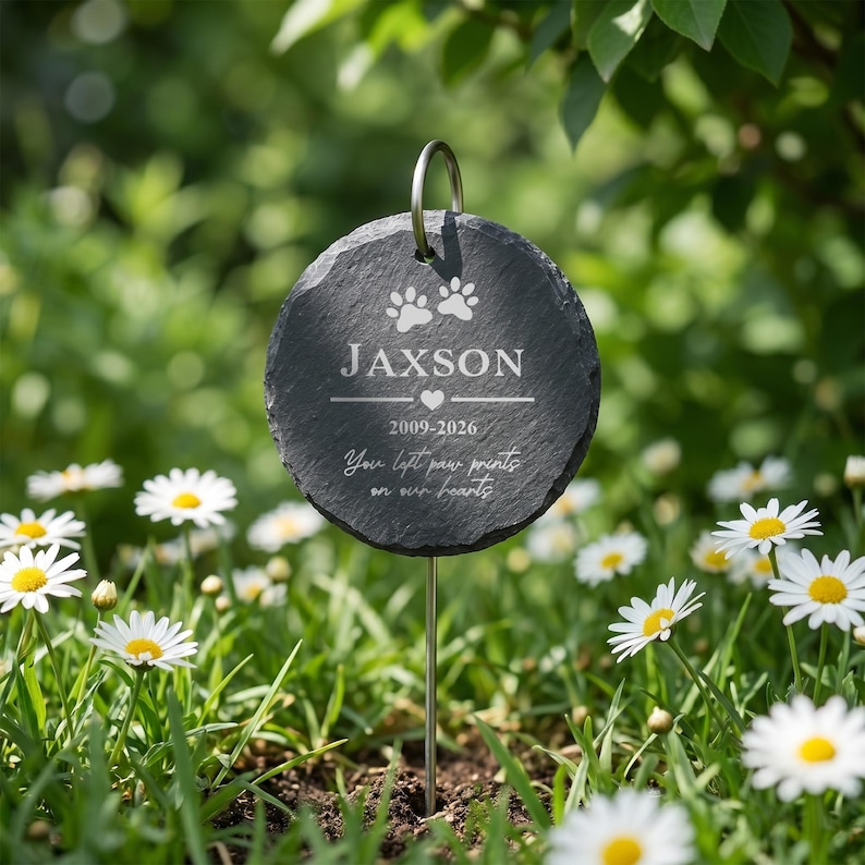 May include: A round, dark gray slate pet memorial stake with the name "Jaxson", paw prints, and dates 2009-2026. The text "You left paw prints on our hearts" is also included. The stake is in a garden with daisies.