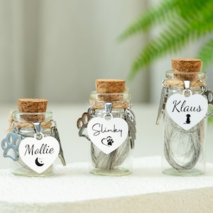 May include: Three glass memorial jars with cork stoppers, each containing pet hair. Each jar has a heart-shaped tag with a pet's name and paw print charm. The names are Mollie, Slinky, and Klaus.