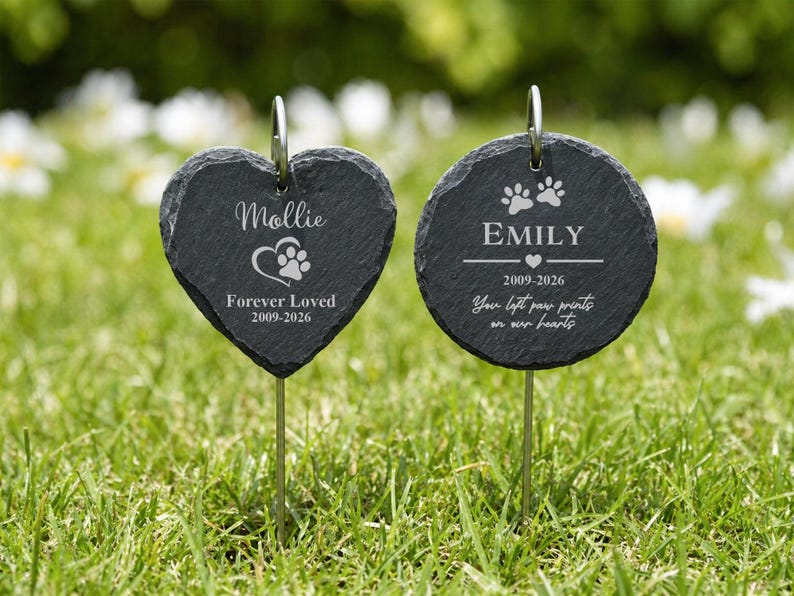 May include: Two black slate pet memorial garden stakes. One is heart-shaped with the name "Mollie", a paw print, and the text "Forever Loved 2009-2026". The other is round with "Emily", paw prints, and the text "You left paw prints on our hearts".