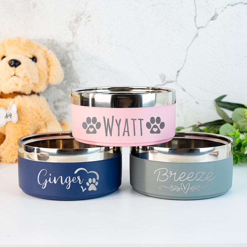 Puede incluir: Tres cuencos para mascotas de acero inoxidable con exteriores de colores. Uno es rosa con "Wyatt" y huellas de patas, otro es azul marino con "Ginger" y una huella de pata, y el tercero es gris con "Breeze" y corazones.