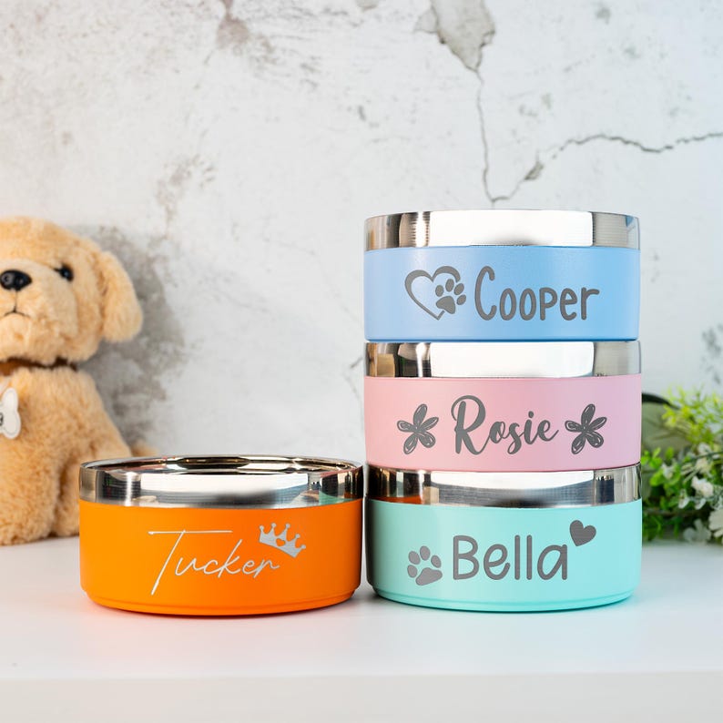 Puede incluir: Pila de cuatro cuencos coloridos para mascotas con bordes de acero inoxidable. Los cuencos son de color naranja, rosa, azul claro y turquesa. Cada cuenco tiene grabado el nombre de una mascota, junto con una huella de pata, un coraz&oacute;n o un dise&ntilde;o de corona. Un perro de peluche est&aacute; en el fondo.