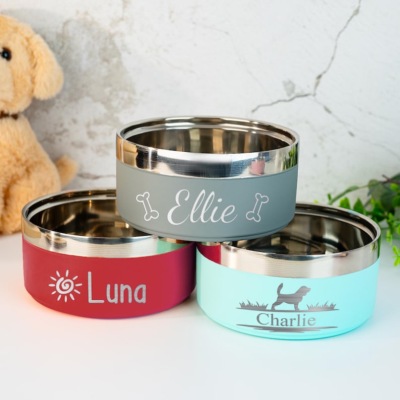 Puede incluir: Tres cuencos para mascotas de acero inoxidable con exteriores de colores. Uno es rojo con "Luna" y un gr&aacute;fico de sol, otro es gris con "Ellie" y gr&aacute;ficos de huesos, y el tercero es azul claro con "Charlie" y una silueta de perro.