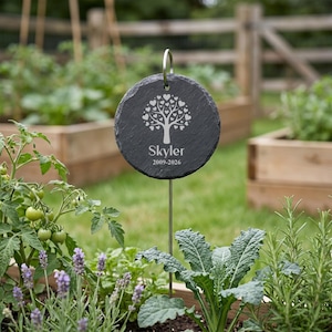 May include: A round, dark gray slate garden marker with a silver metal stake. The marker features a tree design made of hearts and the name "Skyler" with the dates 2009-2026. The marker is placed in a garden bed with various plants.