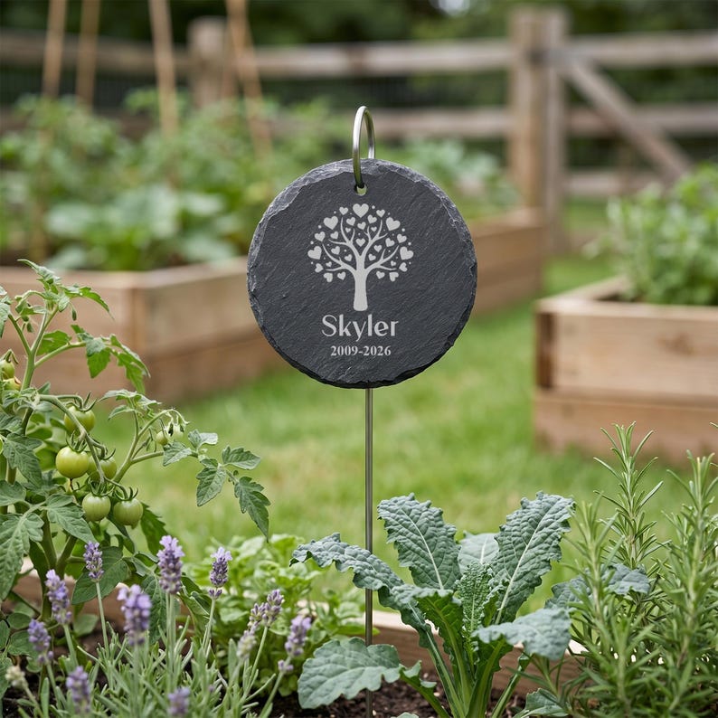 May include: A round, dark gray slate garden marker with a silver metal stake. The marker features a tree design made of hearts and the name "Skyler" with the dates 2009-2026. The marker is placed in a garden bed with various plants.
