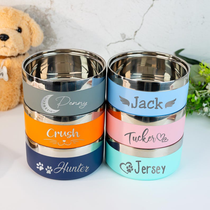 Puede incluir: Una pila de seis cuencos para mascotas de acero inoxidable con bandas de colores gris, naranja y azul. Cada cuenco est&aacute; grabado con el nombre de una mascota y un icono decorativo. Los cuencos son para comida o agua.