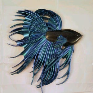 May include: A wall-mounted metal sculpture of a betta fish. The fish has a dark body and iridescent blue and green fins. The fins are detailed with a flowing, feathery design, creating a sense of movement and elegance. The sculpture is mounted on a white background.
