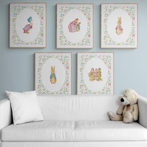 May include: Five framed watercolour prints showcasing illustrations of storybook characters. Each print is encircled by a floral border. The characters include a duck, rabbits, and a scene with a fence and watering can. The prints are displayed above a white sofa.