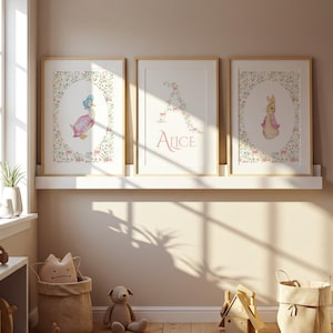 May include: Three framed watercolor prints featuring children's illustrations. One print depicts a duck in a pink dress and blue bonnet, another displays the name "Alice", and the third shows a rabbit in a pink jacket. All are surrounded by floral borders.