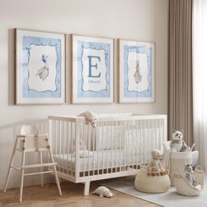 May include: Three framed watercolor prints with light blue polka dot borders and illustrations of a goose, the letter "E" with the name "Edward", and a rabbit. A white crib, high chair, and stuffed animals are in the foreground.