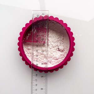 May include: Overhead shot of a fuchsia pink, scalloped-edge circular container filled with white powder. A clear ruler, marked in inches, is placed across the container, showing measurements up to 12 inches. The container is on a white surface.