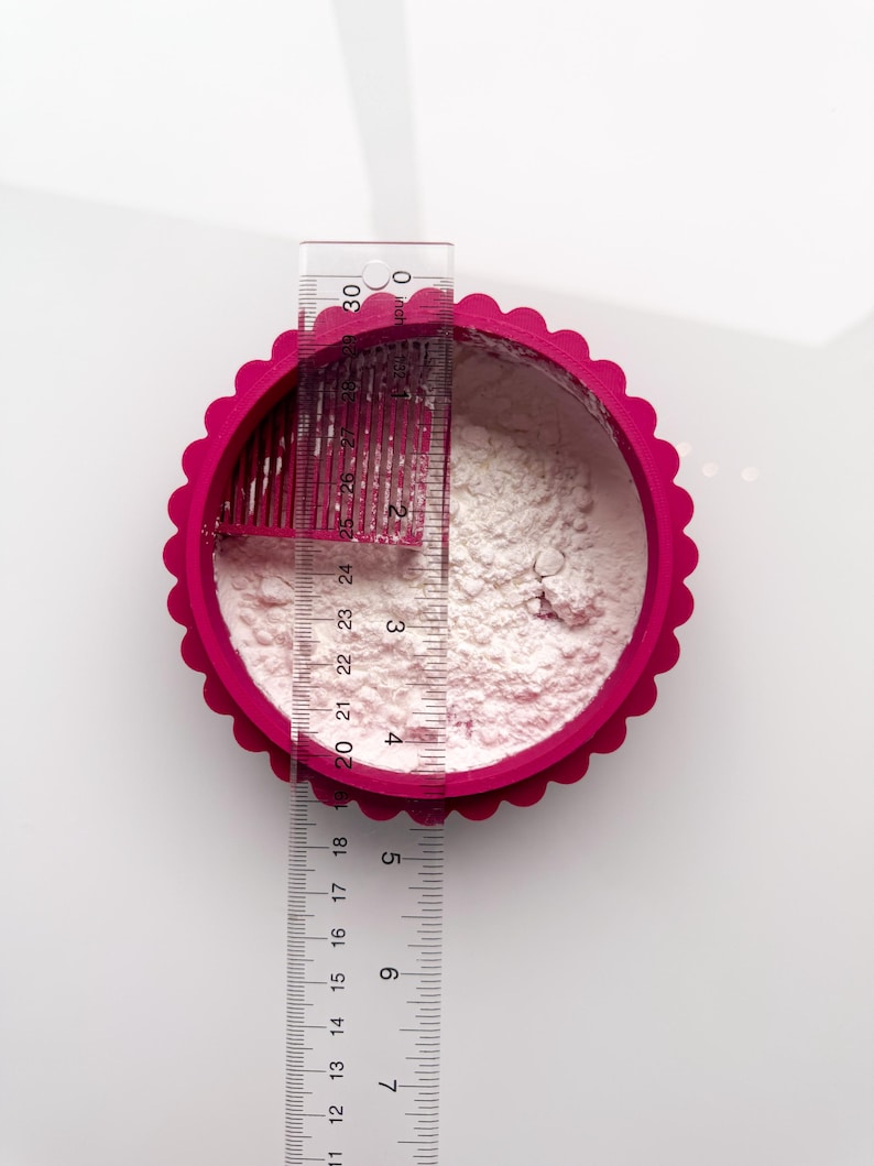 May include: Overhead shot of a fuchsia pink, scalloped-edge circular container filled with white powder. A clear ruler, marked in inches, is placed across the container, showing measurements up to 12 inches. The container is on a white surface.