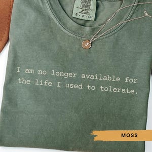 May include: Olive green t-shirt with the text "I am no longer available for the life I used to tolerate." A gold necklace with a pendant rests on the shirt. The shirt is labeled "MOSS".