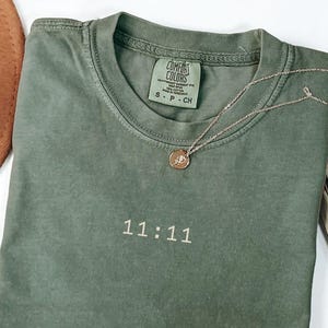 May include: A moss green t-shirt with the embroidered text "11:11" in white. A gold-coloured necklace with a pendant rests on the shirt. The shirt is folded and has a Comfort Colors tag.