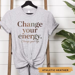 May include: Athletic heather gray t-shirt with the words "Change your energy. Change your life." printed in brown. The shirt is knotted at the waist and hangs on a wooden ladder.
