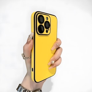 Puede incluir: Una funda de teléfono de color amarillo brillante con un borde negro. La funda tiene un recorte para la cámara con círculos negros. La funda del teléfono se sostiene en una mano con uñas largas y manicuradas y anillos.