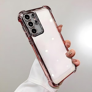 Peut inclure: Coque de téléphone transparente avec un cadre or rose. L'étui a un intérieur blanc et une conception de pare-chocs de protection. L'étui du téléphone est tenu dans une main.