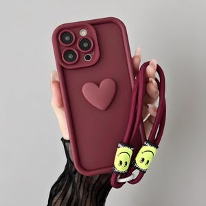May include: A claret-coloured phone case featuring a raised heart. It has a matching claret lanyard with smiley face charms. The case is designed for a phone with multiple camera lenses.