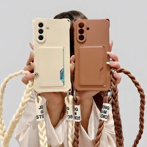 May include: Two phone cases, one cream and one brown, are held up in front of a person. Each case has a card slot and a braided strap. The cream case has a white strap, and the brown case has a brown strap. The cases have a camera module with three lenses.