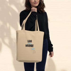 Può includere: Borsa tote beige con testo nero "LAGO STRESA MAGGIORE" e testo azzurro "ITALIA". Un disegno di scooter è sotto il testo. La borsa ha due manici beige.