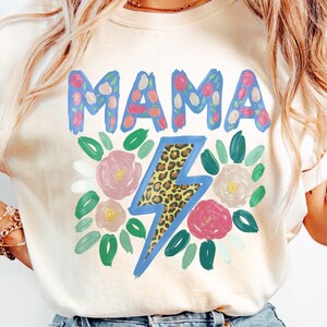 Könnte beinhalten: Cremefarbenes T-Shirt mit dem Wort "MAMA" in blauen, mit Blumen gefüllten Buchstaben. Ein Blitz mit Leopardenmuster ist unter dem Wort zentriert, umgeben von rosa und weißen Blumen und grünen Blättern. Das Shirt hat einen Rundhalsausschnitt.
