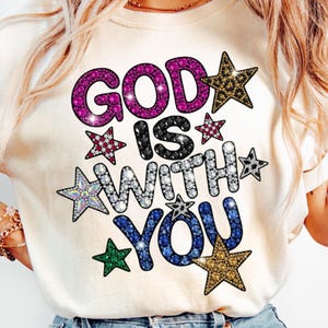 Puede incluir: Camiseta de color crema con la frase "GOD IS WITH YOU" en un diseño brillante con tonos joya. El texto está rodeado de estrellas de varios colores, incluyendo rosa, dorado y azul. Una camiseta gráfica casual e inspiradora.