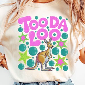 May include: A light beige t-shirt featuring a cartoon kangaroo and the words "Too Da Loo" in pink bubble letters. The design includes teal circles and green and pink stars, creating a playful and colorful graphic.