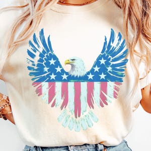 May include: Cream-coloured t-shirt with a patriotic design. The graphic shows a stylised bald eagle with wings in blue, white stars, and red and white stripes, resembling the American flag. The eagle's head is white with a yellow beak.