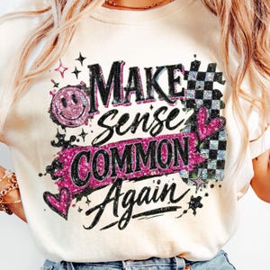Può includere: T-shirt color crema con la scritta "MAKE Sense COMMON Again" in nero, rosa e glitter argento. Il design include una faccina sorridente, cuori e un motivo a scacchi. Un capo casual.