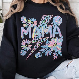 Könnte beinhalten: Schwarzes Sweatshirt mit dem Wort "MAMA" in Pastellfarben, mit einem Blitz und floralen Akzenten. Der Blitz hat ein Leopardenmuster. Die Blumen sind in Rosa, Blau und Gelb gehalten.
