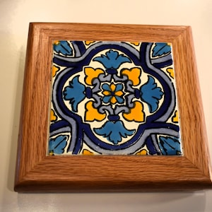 May include: Square wooden framed tile with a colorful floral design. The tile features a central flower with blue and yellow petals, surrounded by a blue and white pattern. The frame is made of light brown wood.
