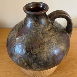 May include: A brown ceramic vase with a mottled glaze and a wooden base. The vase has a rounded body and a single handle.