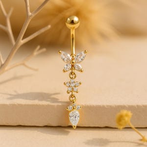 Puede incluir: Anillo de ombligo dorado con un colgante de mariposa, un colgante de flor y un colgante de lágrima. Cada colgante está adornado con piedras de circonita cúbica transparentes.