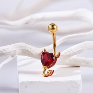 Puede incluir: Anillo de ombligo dorado con una gema roja en forma de corazón y un diseño de cola de diablo.