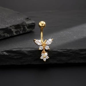 Puede incluir: Anillo para el ombligo de color dorado con un diseño de mariposa y flor. La mariposa tiene dos piedras transparentes en forma de lágrima para las alas y un grupo de pequeñas piedras para el cuerpo. Una pequeña flor con piedras transparentes cuelga debajo.
