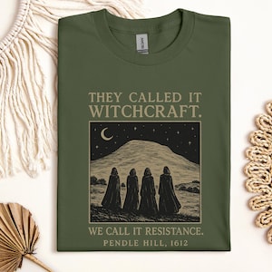 May include: Olive green t-shirt with the text "THEY CALLED IT WITCHCRAFT. WE CALL IT RESISTANCE. PENDLE HILL, 1612." The graphic depicts figures in cloaks standing before a mountain under a crescent moon and stars. The shirt has a crew neck.