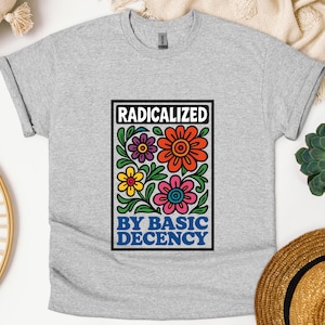 May include: A light grey t-shirt featuring a vibrant floral design within a black rectangle. The text reads "RADICALIZED" above and "BY BASIC DECENCY" below. The flowers are in shades of orange, pink, yellow, and purple.